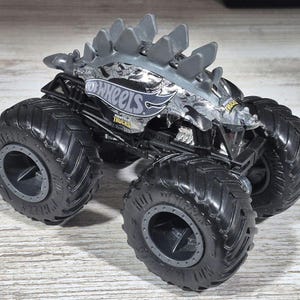 May include: A grey and black Hot Wheels monster truck toy with large, black tyres. The truck features a dinosaur-like design on the top and the Hot Wheels logo on the side. The tyres are thick and have a deep tread.