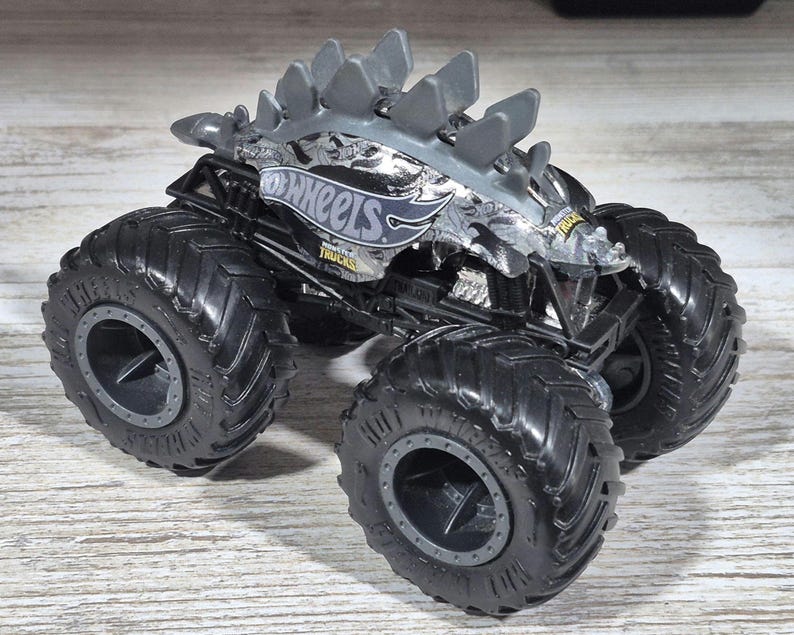 May include: A grey and black Hot Wheels monster truck toy with large, black tyres. The truck features a dinosaur-like design on the top and the Hot Wheels logo on the side. The tyres are thick and have a deep tread.