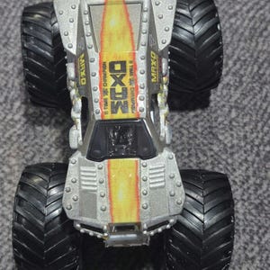 May include: A silver and yellow MAX-D monster truck toy with large black tyres. The truck has the text "MAX-D" and "11 TIME CHAMPION" printed on it. The truck has a detailed design with rivets and a flame-like pattern.