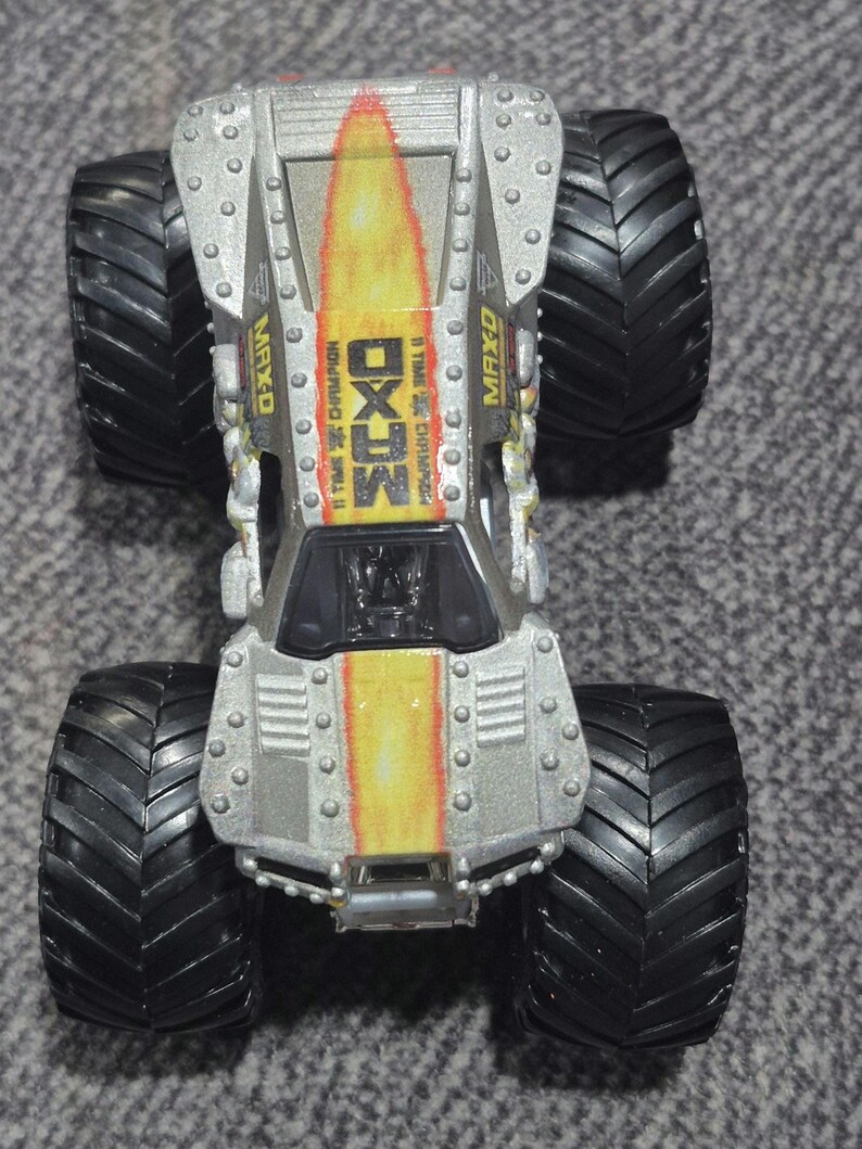 May include: A silver and yellow MAX-D monster truck toy with large black tyres. The truck has the text "MAX-D" and "11 TIME CHAMPION" printed on it. The truck has a detailed design with rivets and a flame-like pattern.