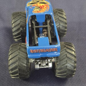May include: A blue monster truck toy with large black tires. The truck features a skull and flame design on the roof and the word "INSTIGATOR" on the side. The toy is made of metal and plastic.