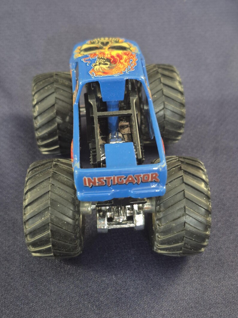 May include: A blue monster truck toy with large black tires. The truck features a skull and flame design on the roof and the word "INSTIGATOR" on the side. The toy is made of metal and plastic.