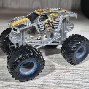 May include: A silver and yellow MAX-D monster truck toy with large black tyres and blue rims. The truck features a detailed design with the MAX-D logo and a graphic of a skull with mechanical arms.