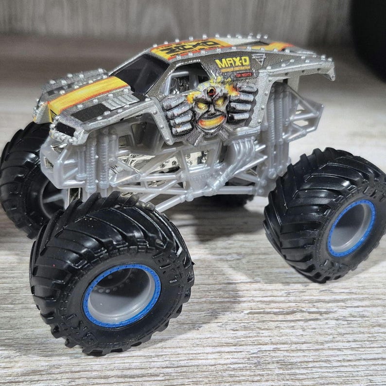 May include: A silver and yellow MAX-D monster truck toy with large black tyres and blue rims. The truck features a detailed design with the MAX-D logo and a graphic of a skull with mechanical arms.