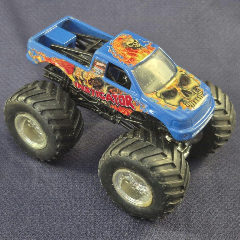 May include: A blue monster truck toy with large black tires and silver rims. The truck features a skull and flame design on the hood and sides, with the word "INSTIGATOR" in red. The truck is set against a blue background.