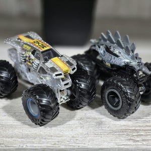 May include: Two toy monster trucks with large black tyres. One truck is clear with yellow and black accents and the text "MXO". The other truck is black with grey spikes on top and the text "Hot Wheels".