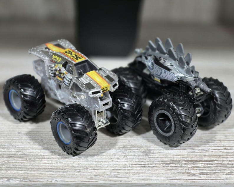 May include: Two toy monster trucks with large black tyres. One truck is clear with yellow and black accents and the text "MXO". The other truck is black with grey spikes on top and the text "Hot Wheels".