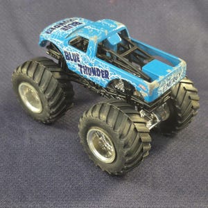 May include: A blue monster truck toy with the words "BLUE THUNDER" and lightning bolt graphics. The truck has large black tires and silver rims. The toy truck is a miniature replica of a monster truck.