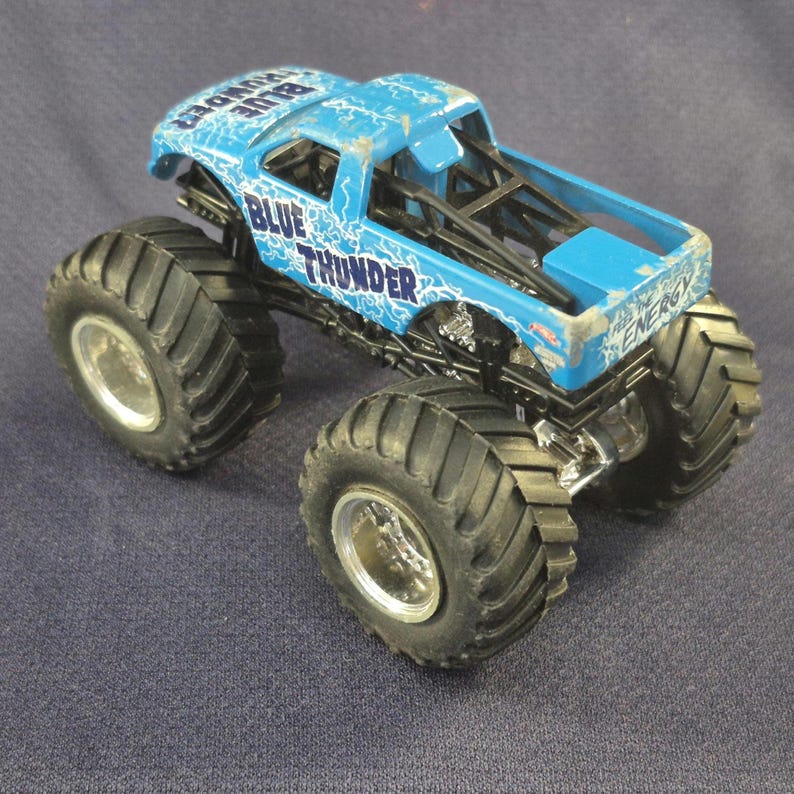 May include: A blue monster truck toy with the words "BLUE THUNDER" and lightning bolt graphics. The truck has large black tires and silver rims. The toy truck is a miniature replica of a monster truck.