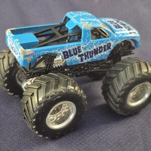 May include: A blue toy monster truck with the words "BLUE THUNDER" and lightning bolt graphics. The truck has large black tires and a black chassis. The toy truck is a Hot Wheels model.