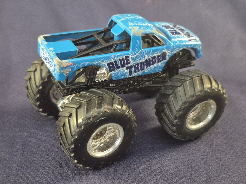 May include: A blue toy monster truck with the words "BLUE THUNDER" and lightning bolt graphics. The truck has large black tires and a black chassis. The toy truck is a Hot Wheels model.
