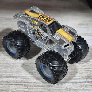 May include: A silver and black toy monster truck with large black tyres and blue rims. The truck has a yellow and orange flame design on the hood and the text "MAX-D" and "OX2" on the body. The toy vehicle is a miniature truck.