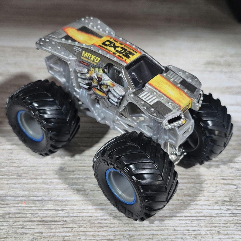 May include: A silver and black toy monster truck with large black tyres and blue rims. The truck has a yellow and orange flame design on the hood and the text "MAX-D" and "OX2" on the body. The toy vehicle is a miniature truck.