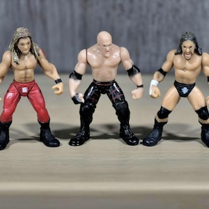 May include: Three small, plastic wrestling action figures. The figures are muscular, with detailed facial features and wrestling attire. One figure wears red pants, another black pants, and the third black trunks. All have black boots and wristbands.