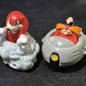 May include: Two plastic character figurines. On the left, a red and white Knuckles figure emerges from a textured white cloud base. On the right, a Dr. Eggman figure with orange mustache and red suit sits inside a gray circular vehicle with a yellow light.