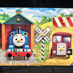 May include: A wooden peg puzzle featuring Thomas the Tank Engine in a blue train shed, a red bus, a white and black rail crossing sign, and a red and white striped barrier. The scene includes green hills, blue skies, and yellow peg handles.