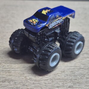 A blue toy monster truck with black wheels and the word "SHOCKER" on the side. The truck has a yellow lightning bolt design on the hood