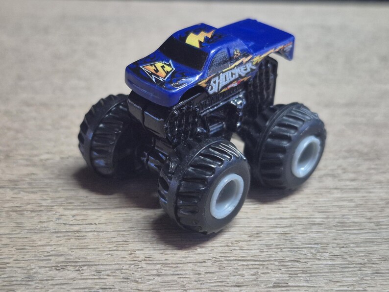 A blue toy monster truck with black wheels and the word "SHOCKER" on the side. The truck has a yellow lightning bolt design on the hood