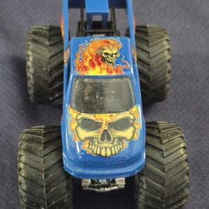 May include: Blue monster truck toy with large black tires. The truck features a skull graphic with flames on the hood and roof. The toy has a roll cage and a clear windshield.