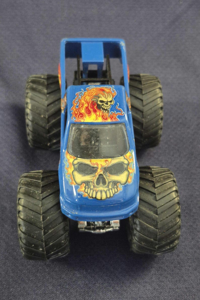 May include: Blue monster truck toy with large black tires. The truck features a skull graphic with flames on the hood and roof. The toy has a roll cage and a clear windshield.