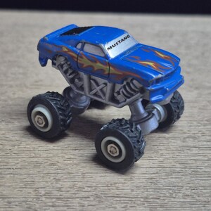 A blue toy monster truck with the word "MUSTANG" on the windshield. The truck has large black tires and a silver chassis. Flames are painted on the hood and sides of the vehicle.