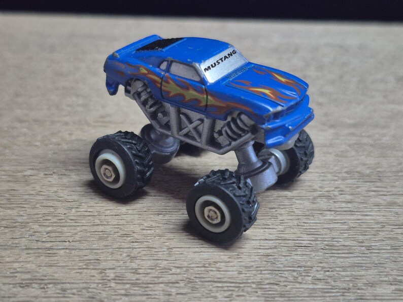 A blue toy monster truck with the word "MUSTANG" on the windshield. The truck has large black tires and a silver chassis. Flames are painted on the hood and sides of the vehicle.