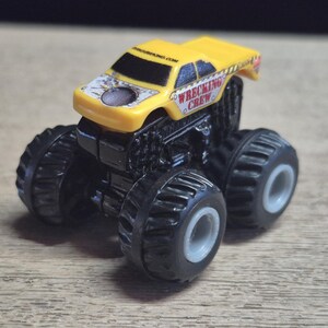 A yellow toy monster truck with black wheels and chassis. The truck has the words "WRECKING CREW" printed on the side and a design on the hood. .