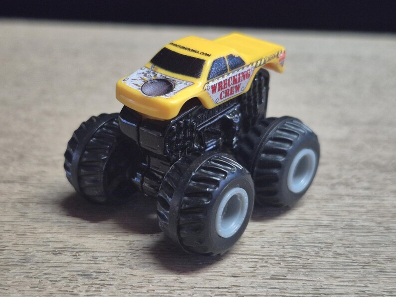 A yellow toy monster truck with black wheels and chassis. The truck has the words "WRECKING CREW" printed on the side and a design on the hood. .
