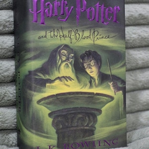 May include: A hardcover book titled "Harry Potter and the Half-Blood Prince" with a green and purple cover. The cover features illustrations of Dumbledore, Harry Potter, and a glowing bowl. The author's name, J.K. Rowling, is at the bottom.