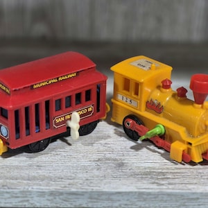 May include: Two toy trains: a red trolley car with "Municipal Railway" and "San Francisco 18" text, and a yellow locomotive with the number "125". Both are plastic and sit on a wooden surface.