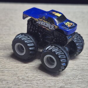 A small, blue and black toy monster truck with large tires. The truck has the word "SHOCKER" in yellow and blue lettering on the side and a lightning bolt graphic. The tires are black with gray rims.