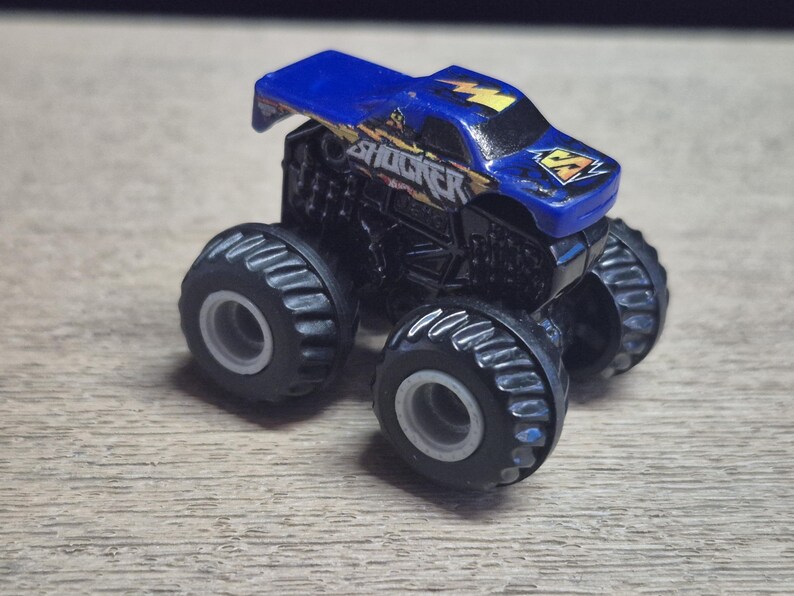A small, blue and black toy monster truck with large tires. The truck has the word "SHOCKER" in yellow and blue lettering on the side and a lightning bolt graphic. The tires are black with gray rims.