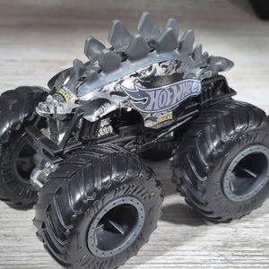 May include: A silver and black Hot Wheels monster truck toy. The toy truck has large, black, textured tyres and a spiked roof design. The Hot Wheels logo is visible on the side of the truck.