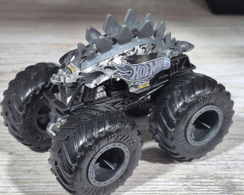 May include: A silver and black Hot Wheels monster truck toy. The toy truck has large, black, textured tyres and a spiked roof design. The Hot Wheels logo is visible on the side of the truck.