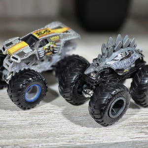 May include: Two toy monster trucks. One is silver with yellow and black accents, the other is grey with a spiky design on top. Both have large black tyres with detailed treads and blue or grey centres.