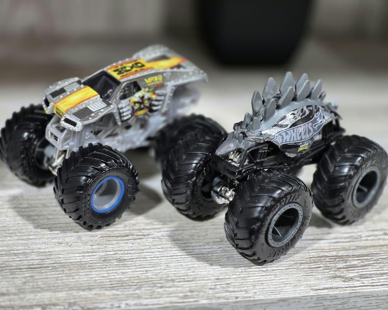 May include: Two toy monster trucks. One is silver with yellow and black accents, the other is grey with a spiky design on top. Both have large black tyres with detailed treads and blue or grey centres.