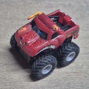 A red monster truck toy with large black tires and a detailed design. The truck features a bull-like face with a gold horn and a flame graphic.