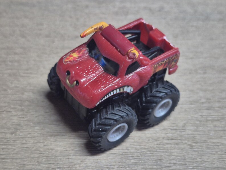 A red monster truck toy with large black tires and a detailed design. The truck features a bull-like face with a gold horn and a flame graphic.
