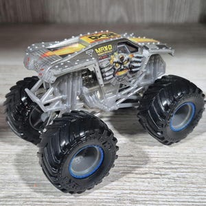 May include: A silver and yellow toy monster truck with the name "MAX-D" printed on the side. The truck has large black tyres with blue accents and a detailed engine design. The body features a graphic of a skull and mechanical arms.