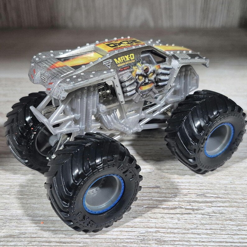 May include: A silver and yellow toy monster truck with the name "MAX-D" printed on the side. The truck has large black tyres with blue accents and a detailed engine design. The body features a graphic of a skull and mechanical arms.