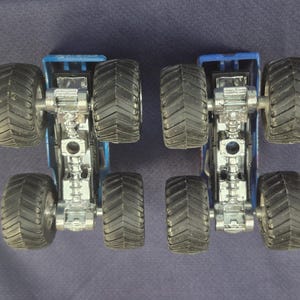 May include: Two blue monster truck toys with large black tires and silver chassis are positioned side-by-side. The trucks are viewed from below, showcasing the intricate details of the undercarriage and tire treads. The background is a solid dark blue.