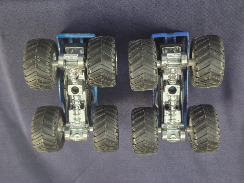 May include: Two blue monster truck toys with large black tires and silver chassis are positioned side-by-side. The trucks are viewed from below, showcasing the intricate details of the undercarriage and tire treads. The background is a solid dark blue.