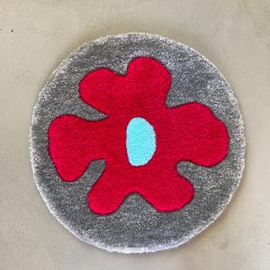 Peut inclure: Un tapis rond gris avec un motif de fleur rouge et un centre bleu clair. Le tapis a un aspect doux et texturé, et la forme de la fleur ressort sur le fond gris. Convient pour une chambre ou un salon.