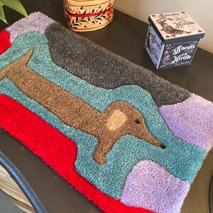 Op de afbeelding: Een kleurrijk, handgemaakt vloerkleed met een bruin teckelontwerp. Het vloerkleed heeft een rode rand, turquoise, grijze en paarse secties. Een hartvormige doos met bloemmotieven en een koekjesdoos zijn ook zichtbaar.