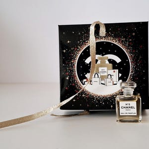 Puede incluir: Un peque&ntilde;o frasco de perfume Chanel No. 5 Eau de Parfum se encuentra frente a una caja de regalo negra con una cinta dorada. La caja presenta un dise&ntilde;o circular con el logo de Chanel e ilustraciones de frascos de perfume y bolsas de compras.