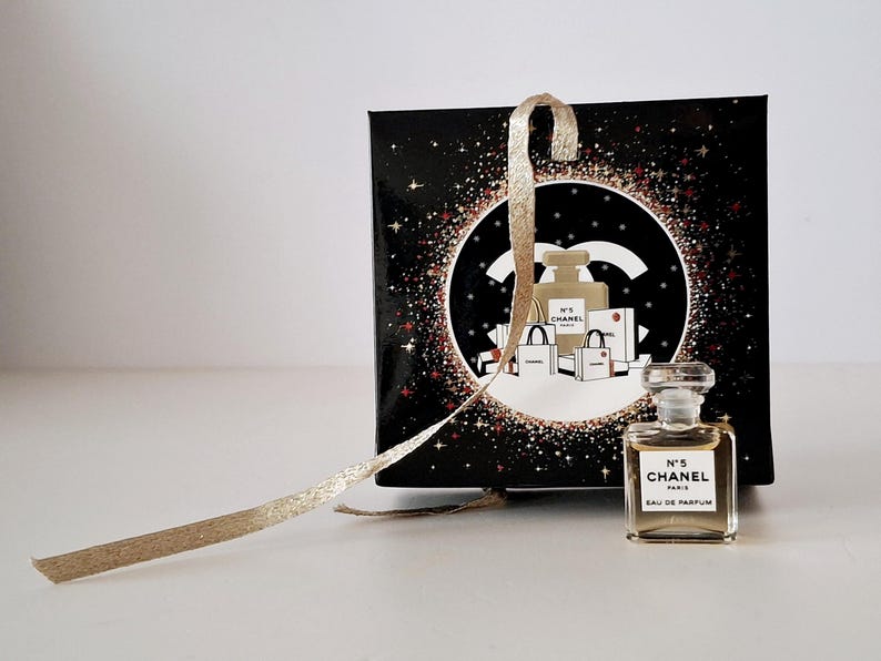 Puede incluir: Un peque&ntilde;o frasco de perfume Chanel No. 5 Eau de Parfum se encuentra frente a una caja de regalo negra con una cinta dorada. La caja presenta un dise&ntilde;o circular con el logo de Chanel e ilustraciones de frascos de perfume y bolsas de compras.