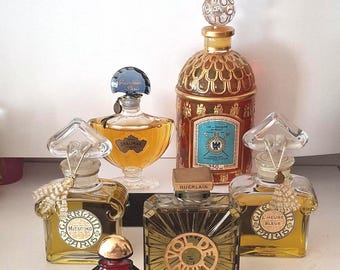 May include: A collection of six Guerlain perfume bottles including Mitsouko, Shalimar, L'Heure Bleue, Vol de Nuit, Samsara, and a large Eau de Cologne bottle. The bottles feature various glass shapes, stoppers, and labels with gold and amber liquid.