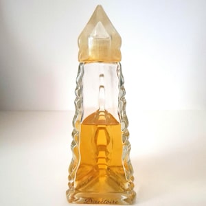 May include: A clear glass perfume bottle with a unique lighthouse shape, featuring a pointed top and a textured, ridged design along the sides. The bottle contains a golden-colored liquid and has the word "Directoire" printed on the base.