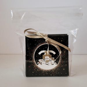 Puede incluir: Una peque&ntilde;a caja negra cuadrada con una cinta dorada, dentro de una bolsa de pl&aacute;stico transparente. La caja presenta un dise&ntilde;o circular con el logo de Chanel e ilustraciones de bolsas de compras. El fondo es negro con purpurina dorada.