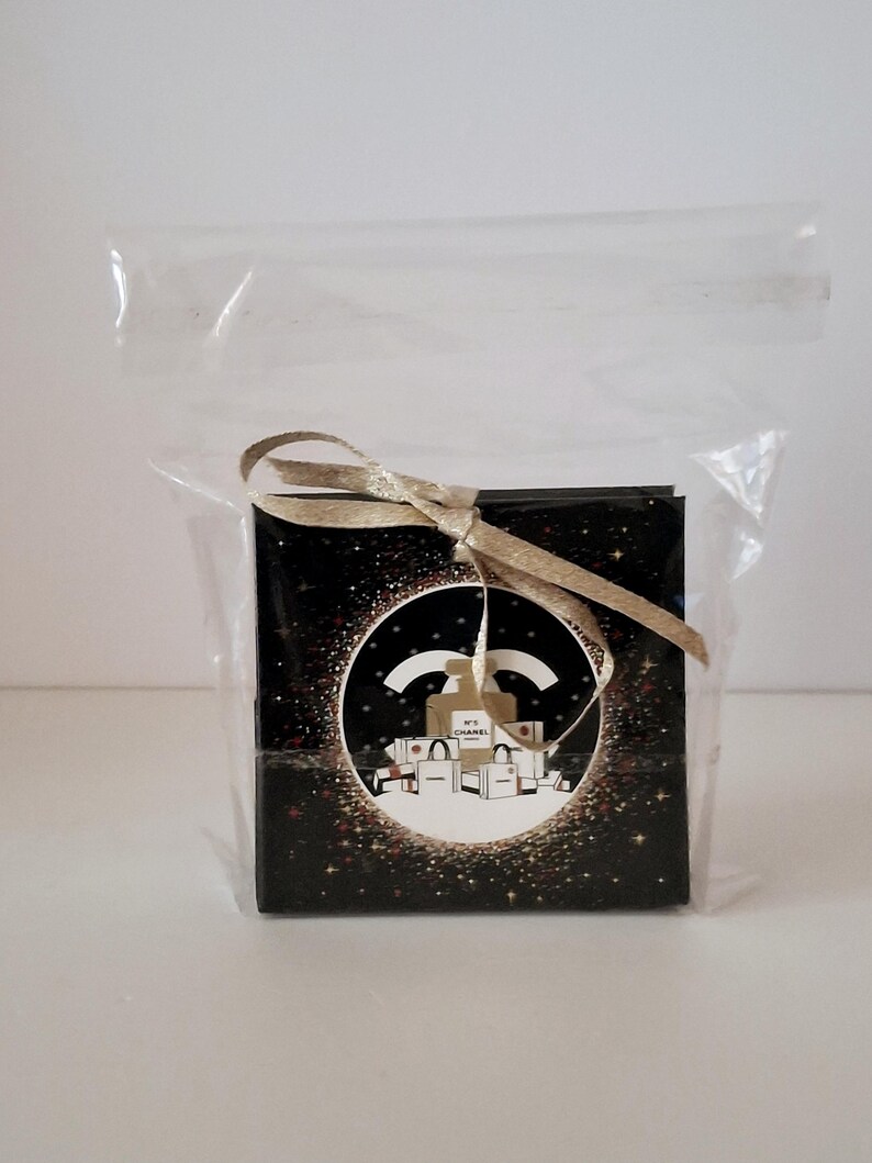 Puede incluir: Una peque&ntilde;a caja negra cuadrada con una cinta dorada, dentro de una bolsa de pl&aacute;stico transparente. La caja presenta un dise&ntilde;o circular con el logo de Chanel e ilustraciones de bolsas de compras. El fondo es negro con purpurina dorada.